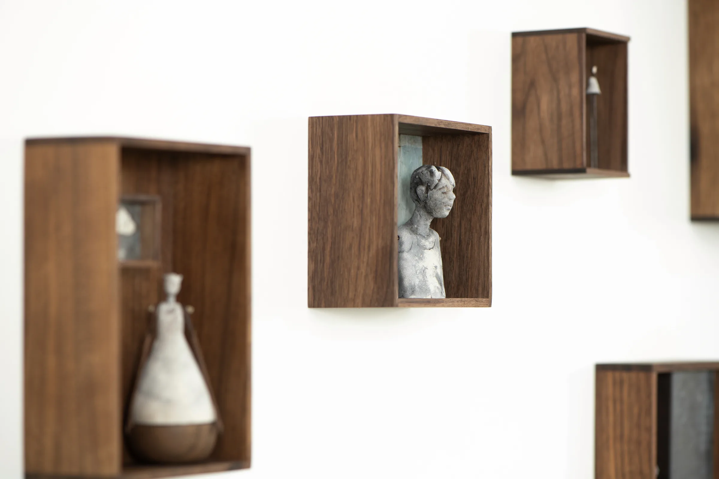 Installation view of Echoes of Home — walnut box sculptures with porcelain figures on the gallery wall